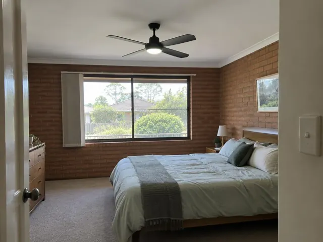 Bedroom with a ceiling fan above a bed, brick walls and a large window in Beresfield